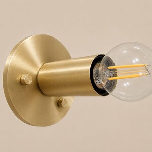 May include: A wall-mounted light fixture featuring a circular brushed brass backplate with two small knurled knobs. A cylindrical brass socket extends from the center, holding a clear glass globe light bulb with visible yellow LED filaments.
