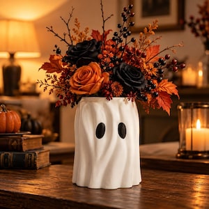 May include: A white ceramic vase shaped like a ghost with two black oval eyes, filled with an autumn floral arrangement featuring orange roses, black roses, dark berries, and orange maple leaves, creating a seasonal decorative display.