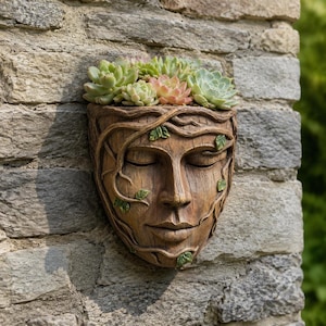 May include: A wall-mounted planter shaped like a human face with closed eyes and a serene expression. The brown, wood-textured surface is adorned with sculpted vines and small green leaves, and it is filled with a cluster of light green succulents.