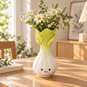 May include: A ceramic vase shaped like a head of napa cabbage with a white base that transitions to a light green ruffled top. The vase features a simple black printed face with two small circular eyes and a curved line for a mouth on the front.