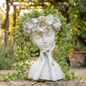 May include: A white head-shaped planter featuring a sculpted face with closed eyes and hands resting under the chin. The head is adorned with a crown of white flowers with gold-painted edges, and long green trailing vines drape over the sides.