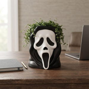 Könnte beinhalten: Ein schwarz-weißer Übertopf in Form einer Geistermaske mit langem, schreiendem Mund und hohlen Augen. Der Übertopf ist mit einer rankenden Grünpflanze gefüllt, die über die oberen Ränder der schwarzen, kapuzenartigen Struktur des Gefäßes hinausragt.