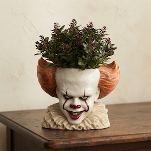 Peut inclure: Un pot de fleurs décoratif en forme de tête de clown avec un visage blanc pâle, des cheveux roux et un col à volants. Le clown porte un maquillage rouge foncé autour des yeux et de la bouche, et le sommet de la tête contient une petite plante verte.