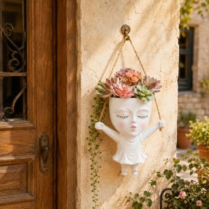 Puede incluir: Una maceta de pared blanca con forma de persona con los ojos cerrados y boca pequeña, que lleva un vestido. La maceta está suspendida por una cuerda color canela de un gancho metálico y contiene suculentas verdes y rosas con enredaderas.