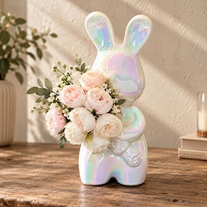 Może przedstawiać: Ceramiczna figurka królika z opalizującym, perłowym wykończeniem, trzymająca bukiet sztucznych jasnoróżowych piwonii i drobnych białych kwiatów. Królik ma długie uszy oraz gładką, odbijającą światło powierzchnię.