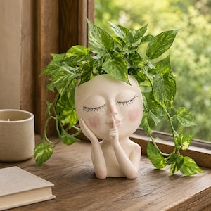 May include: A cream-coloured head-shaped planter featuring a face with closed eyes, pink cheeks, and a finger held to the lips in a shushing gesture. The planter is filled with a trailing green and yellow variegated pothos plant.