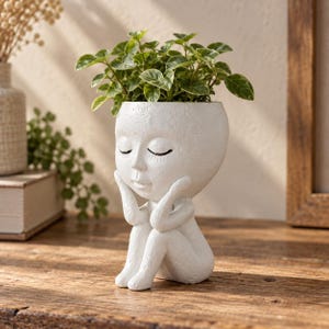 May include: A white ceramic planter shaped like a seated figure with hands resting on the cheeks. The figure has closed eyes and a smooth, textured surface. A small green plant with variegated leaves grows from the top of the head of the figure.