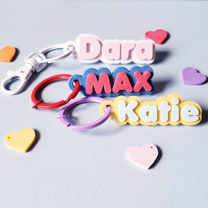 Peut inclure: Trois porte-clés personnalisés avec les noms "Dara", "Max" et "Katie" dans des designs colorés et superposés. Chaque porte-clés a une palette de couleurs différente et est accompagné d'ornements en forme de cœur.