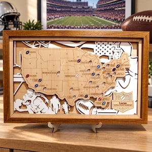 Puede incluir: Una pieza de arte mural con marco de madera rectangular que presenta un mapa en capas de los Estados Unidos con recortes para estadios de fútbol americano. El mapa incluye marcadores con forma de balón y una placa que dice Lucas Football Stadium Tracker Map.