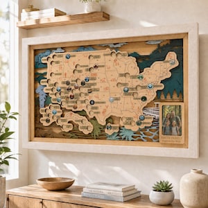 May include: A wooden wall map of the United States featuring layered cutouts of mountains and forests in blue and green tones. The map includes circular slots for tokens and a rectangular photo frame on the right with the text Smith Family Explore the Wild.