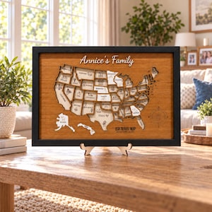 Könnte beinhalten: Eine gerahmte USA-Reisekarte aus Holz mit schwarzem Rand und naturfarbenem Hintergrund. Die Karte zeigt lasergeschnittene Staatsumrisse mit eingravierten Symbolen sowie den Text Annice's Family, USA TRAVEL MAP und ADVENTURE AWAITS.