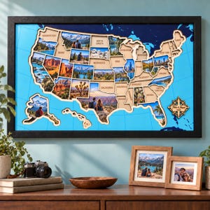 May include: A framed wall map of the United States featuring a blue background with grid lines and wooden state cutouts. Each state contains a photographic print of landscapes or people. A compass rose in the corner displays the text Pattie & Magali.