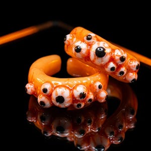 May include: Two orange rings featuring multiple raised, realistic-looking eyeballs with white sclera, red veins, and black pupils. The rings have a glossy, smooth finish and a circular, open-band design, reflecting clearly on a dark surface.