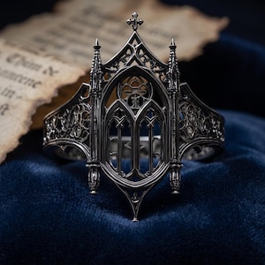 May include: A dark silver-toned metal ring featuring an intricate architectural design reminiscent of a cathedral window. The band displays pointed arches, slender spires, and delicate openwork tracery, culminating in a small cross at the top center.