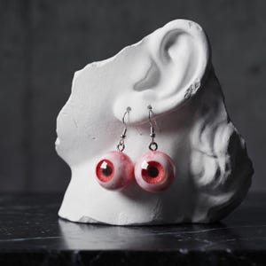 Op de afbeelding: Een paar hangende oorbellen met bolvormige bedels die lijken op realistische menselijke oogbollen. Elke bedel heeft een witte sclera met rode aders en een felrode iris met een zwarte pupil, bevestigd aan zilverkleurige metalen haakjes.