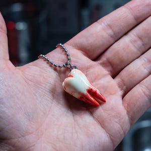 Puede incluir: Un colgante con forma de muela humana con un acabado brillante en color blanco roto y manchas realistas de color rojo intenso en las raíces, suspendido de una cadena de bolas de metal plateado sujeta a una pequeña anilla metálica superior.
