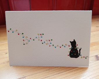 May include: A rectangular white greeting card featuring a black silhouette of a cat sitting and tangled in a string of colorful Christmas lights. The lights are depicted as small red, blue, yellow, and green dots connected by a thin black line.