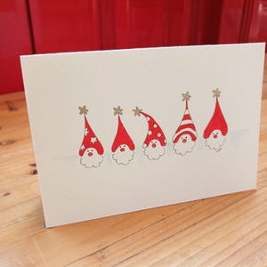 May include: A white greeting card featuring a horizontal row of five illustrated gnomes. Each gnome wears a red hat with a gold star on top, featuring various patterns including white stars, polka dots, and stripes, with white beards and red noses.