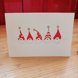 May include: A white greeting card featuring a hand-drawn illustration of five gnomes in a row. Each gnome wears a red hat with unique patterns including stars, polka dots, and stripes, topped with a small gold star. They have white beards.
