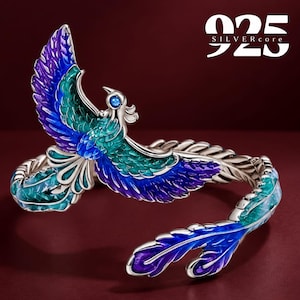 May include: A silver cuff bracelet featuring a phoenix design with blue and green enamel detailing. The bird has a small blue stone for an eye. The text 925 SILVERcore is visible in the upper right corner. The metal band has a textured finish.