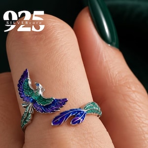May include: A silver open ring featuring an enameled bird design with vibrant purple and green wings and a long tail. The ring displays the text 925 SILVERcore in the upper left corner, indicating the metal composition of the jewelry piece.
