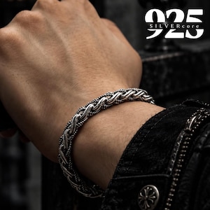 Puede incluir: Una pulsera de plata con un diseño complejo de cadena trenzada similar a una cuerda con detalles oxidados en los huecos. El texto 925 SILVERcore es visible en la esquina superior derecha de la imagen, indicando la composición del material.