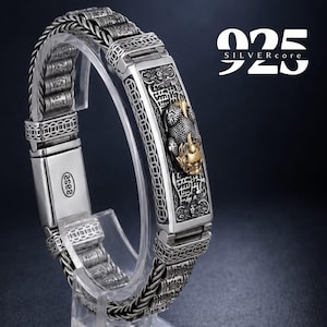 Puede incluir: Una pulsera de plata con una placa rectangular central que presenta una criatura mítica en relieve en tonos oro y plata, rodeada de eslabones con motivos de monedas y segmentos cilíndricos. El cierre tiene el sello 925 y el texto 925 SILVERCORE.