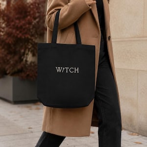 May include: A black canvas tote bag featuring the word WITCH printed in white capital letters on the front, with a small star symbol replacing the letter I. The bag has two black shoulder straps and a rectangular shape with a flat bottom.