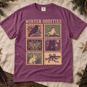 May include: A plum-coloured short-sleeve t-shirt featuring the text WINTER ODDITIES above six rectangular stamp-style illustrations depicting a crow, a ghost near a lighthouse, a moth, a black cat, a rabbit, and a fish tied with a ribbon.