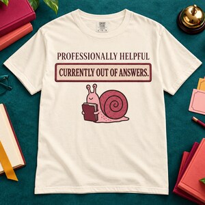 May include: A cream-colored short-sleeve t-shirt featuring a graphic of a pink snail holding a book. Above the snail, the text reads: PROFESSIONALLY HELPFUL CURRENTLY OUT OF ANSWERS. The shirt has a crew neckline and a standard fit.