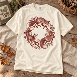 May include: An off-white short-sleeve t-shirt featuring a circular graphic print in dark red ink. The design depicts three foxes arranged in a ring, surrounded by a detailed pattern of blackberry vines, leaves, and berries.