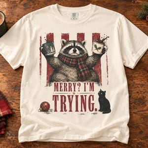 Puede incluir: Una camiseta de manga corta color hueso con un gráfico de un mapache que lleva una bufanda de cuadros rojos y negros y sostiene dos tazas. Debajo del mapache, el texto dice MERRY? I'M TRYING. También aparecen un gato negro y un adorno rojo.