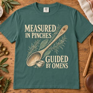 Puede incluir: Una camiseta de manga corta en color verde azulado oscuro con un gráfico de una cuchara de madera, un cuenco pequeño y hierbas. El texto "MEASURED IN PINCHES" y "GUIDED BY OMENS" aparece en color crema. Incluye un símbolo de ojo.