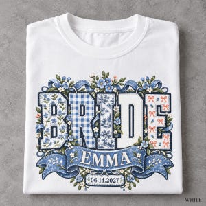 以下が含まれることがあります： 白いTシャツに、青と白の様々な模様で埋め尽くされた大きなブロック体の「BRIDE」という文字が描かれています。その下には、青いリボンのバナーがあり、名前の「EMMA」と日付「2027年6月14日」が記され、青い花々で囲まれています。