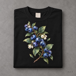 Op de afbeelding: Een zwart t-shirt met ronde hals voorzien van een gedetailleerde botanische illustratie van een bosbessentak. De afbeelding toont trossen ronde, diepblauwe bessen, groene bladeren en kleine witte klokvormige bloemen op de borst.