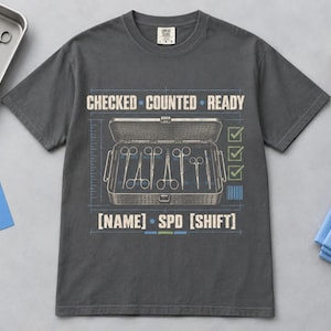 Könnte beinhalten: Ein anthrazitfarbenes T-Shirt mit einer Grafik eines offenen chirurgischen Instrumententabletts. Der Text lautet CHECKED COUNTED READY und [NAME] SPD [SHIFT] mit grünen Häkchen. Das Design enthält blaue Gitterlinien und technische Details.
