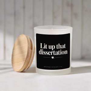 May include: A frosted glass candle in a jar with a bamboo lid. The black label features the white text "Lit up that dissertation" above a graduation cap icon and the text "100% SOY WAX • COTTON WICK • 7 OZ". The candle has a single cotton wick.