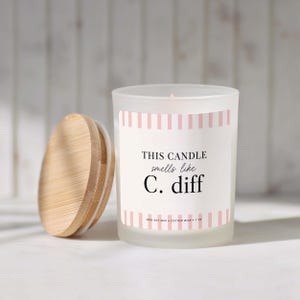 May include: A frosted glass candle in a jar with a bamboo lid. The label features pink vertical stripes and black text that reads: THIS CANDLE smells like C. diff. The label also includes the text 100% SOY WAX • COTTON WICK • 7 OZ.