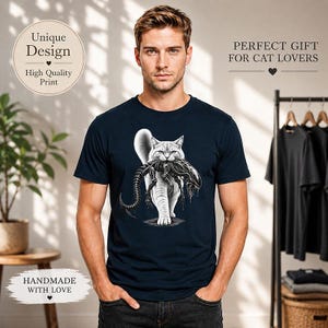 Könnte beinhalten: Ein dunkelblaues T-Shirt mit einem Grafikdruck einer weißen Katze, die vorwärts läuft und ein schwarzes Xenomorph-Wesen im Maul trägt. Die Katze hat eine detaillierte Fellstruktur und das Wesen hat einen langen Schwanz und Körper.