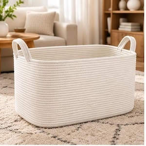May include: A rectangular storage basket constructed from off-white coiled cotton rope. The basket features rounded corners and two integrated rope handles on the short sides. The exterior surface displays a continuous horizontal ribbed texture.