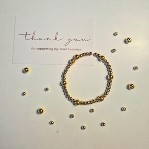 May include: A circular beaded bracelet featuring alternating small and large metallic gold-toned beads. A white rectangular card is positioned behind the bracelet with the text "thank you for supporting my small business" printed in brown.
