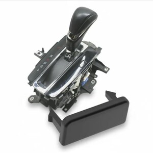 May include: A black automatic transmission gear shifter assembly featuring a chrome-trimmed base with gear indicators labeled P, R, N, D, S, and L. The unit includes a separate black rectangular plastic cover piece positioned in the foreground.