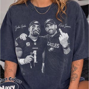 Pode incluir: Uma t-shirt de manga curta azul-marinho com uma impressão gráfica a preto e branco de Jalen Hurts e Nick Sirianni. A impressão inclui o texto Jalen Hurts e Nick Sirianni, com o logótipo Eagles e o número 1 visível na camisola.