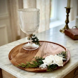 May include: A clear, ribbed glass goblet sits on a round wooden tray. The goblet stem is wrapped in twine with a small green sprig and cross charm. The wooden tray is decorated with two white artificial roses and green pine-like foliage.