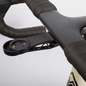 以下が含まれることがあります： マットブラックの自転車用ハンドルステム。黒いバーテープが巻かれ、前方にはサイクルコンピューター用マウントが取り付けられています。ステム上部には円形のロゴが刻印され、マウントの下部にはトラス構造のデザインが施されています。