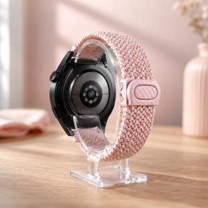 May include: A black smartwatch with a circular sensor array on the back, mounted on a clear acrylic stand. The watch features a braided, textured strap in a light pink and white pattern, secured with a matching pink adjustable sliding buckle.