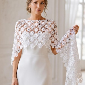 Pode incluir: Um xale de renda branca com um padrão floral intrincado e repetitivo com bordas recortadas. O tecido delicado, semitransparente e estilo crochê, drapeia sobre os ombros e braços, usado sobre um vestido branco simples, liso e sem mangas.