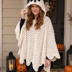 May include: A cream-colored crochet poncho featuring a hood with a ghost face design, including two black oval eyes and a small black circular mouth. The garment has a scalloped hemline and an open-weave texture throughout the fabric.