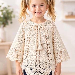 Puede incluir: Un poncho de ganchillo color crema con un intrincado patrón calado, mangas anchas acampanadas y un dobladillo festoneado. La prenda incluye un cuello redondo con un cordón central terminado con dos borlas decorativas a juego.