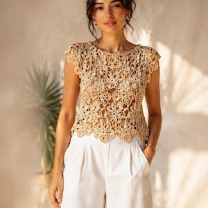Puede incluir: Un top de ganchillo beige con un patrón floral repetitivo, mangas cortas y un dobladillo festoneado, combinado con pantalones blancos de pierna ancha. El top tiene un diseño calado con intrincados motivos florales circulares en toda la prenda.